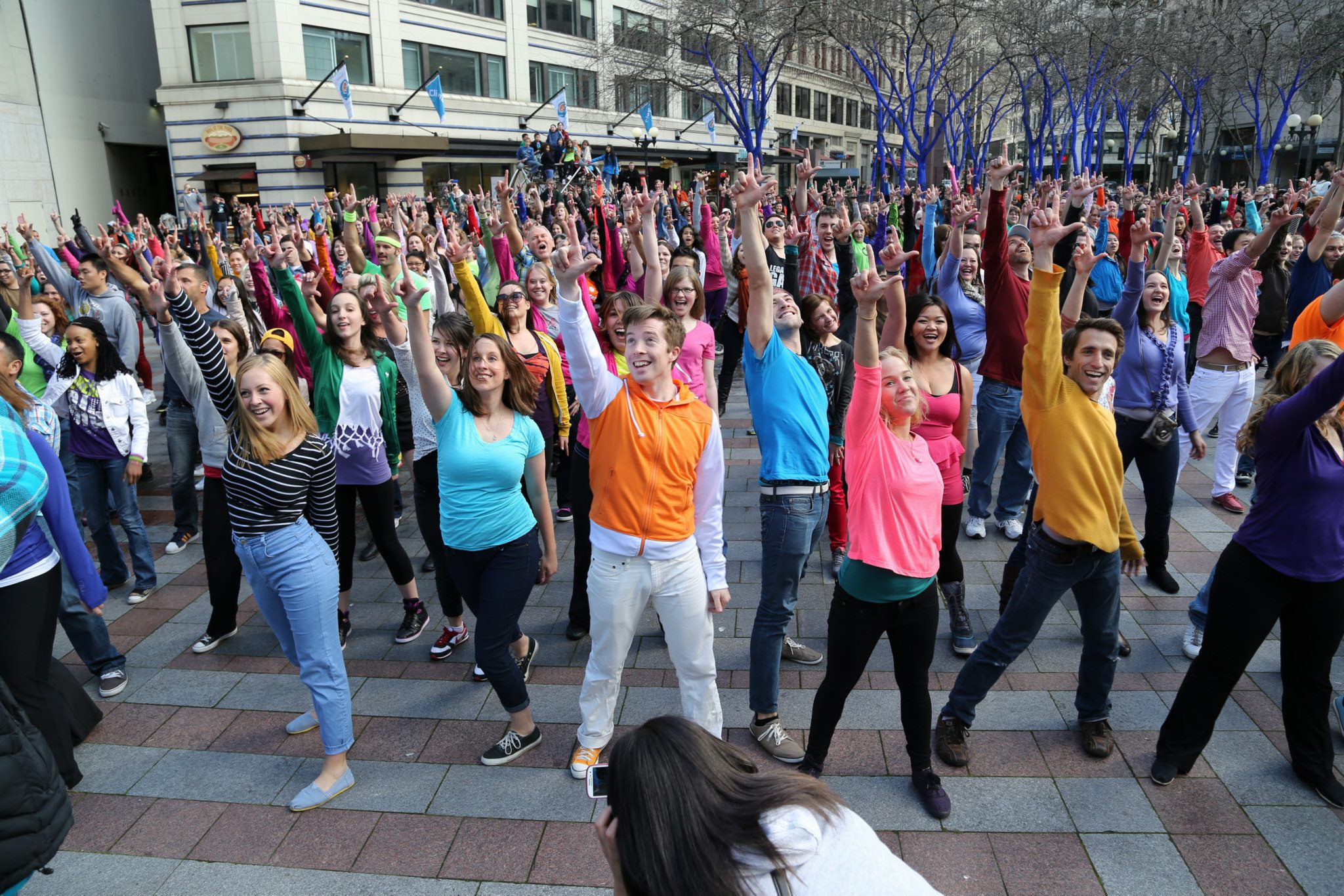 A flash mob crowd of dancers holding 'L' fingers in the air