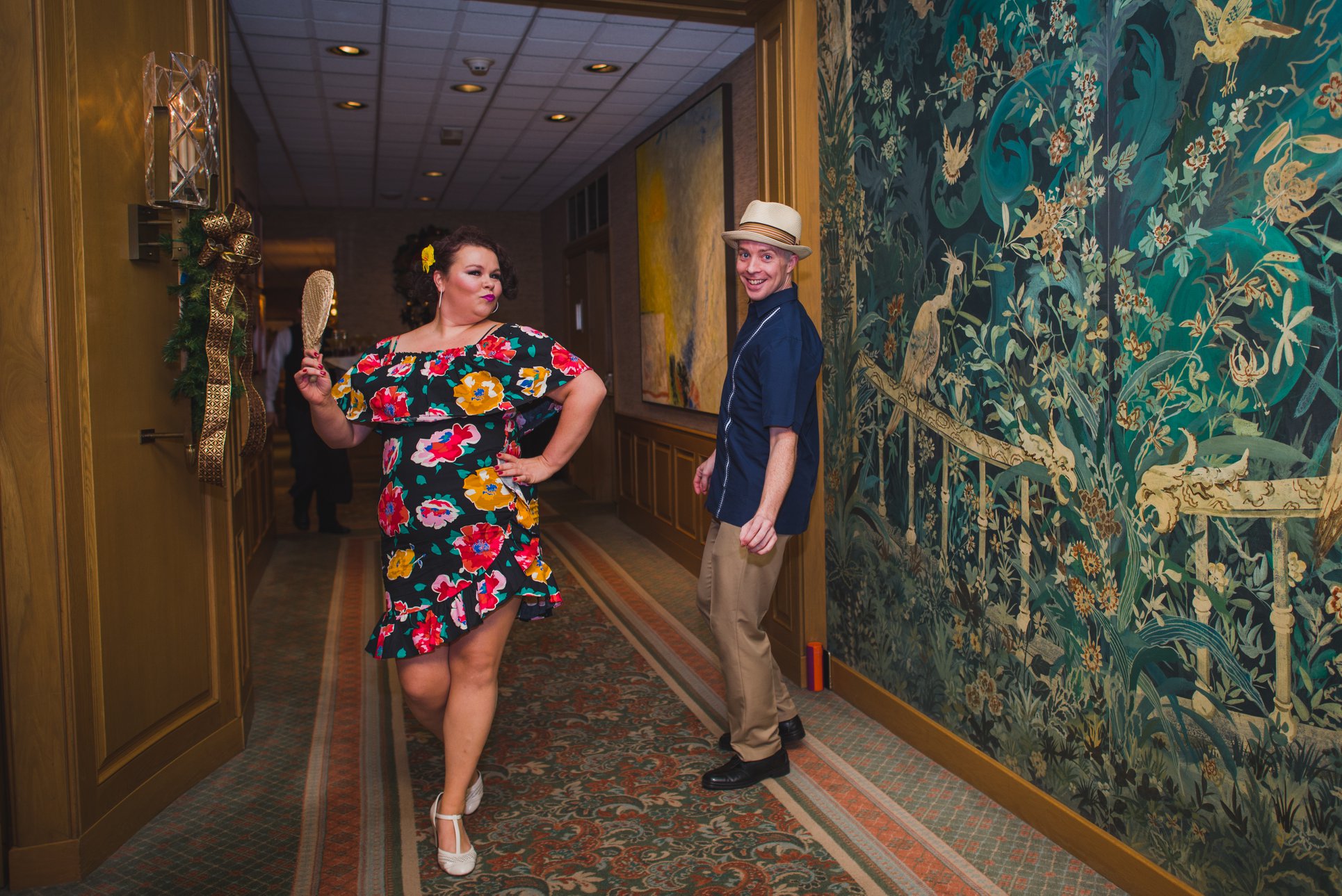 A woman in a flowery dress holding a fan with a sultry pose standing next to a smiling man in a hat.
